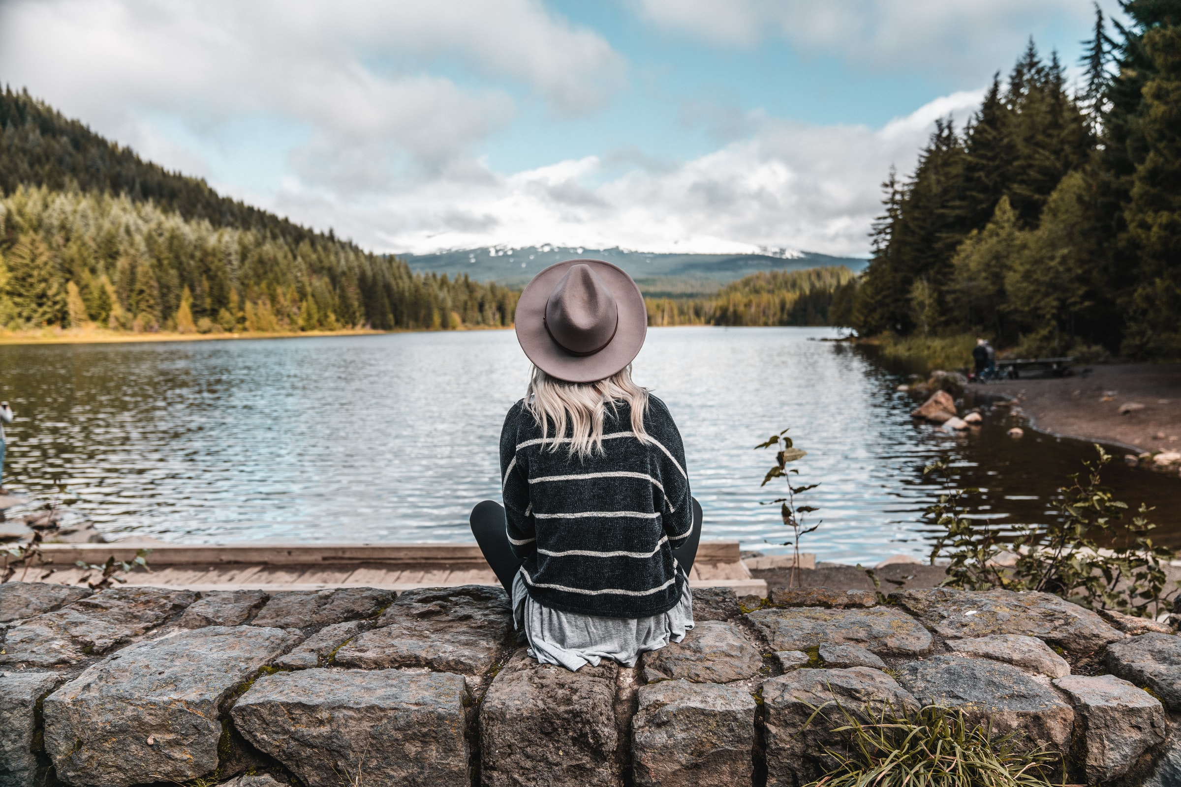 Woman with a hat sitting on a rock wall looking over a lake